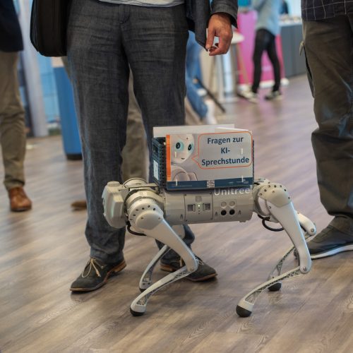 Ein Roboterhund läuft auf vier Beinen herum. Auf ihm drauf klebt ein Schild mit der Aufschrift "Fragen zur KI_Sprechstunde"