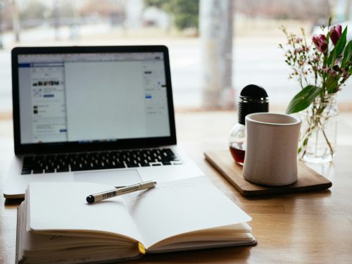 Ein aufgeklappter und eingeschalteter Laptop, davor ein aufgschlagenes Notizbuch mit Stift, daneben eine Kaffeetasse und eine Blumenvase