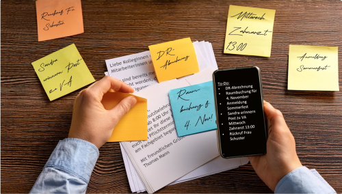 Auf einem Schreibtisch liegen bedruckte weiße Blätter und bunte Klebezettel mit handschriftlichen Notizen. EIne Person hält ein Smartphone darüber. Die Informtationen stehen auch alle auf dem Smartphone.